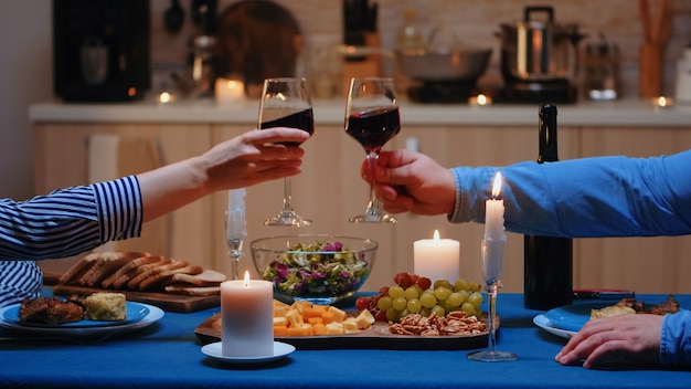 Happy cheerful young couple dining together and clink red wine glasses in the cozy kitchen. Happy caucasian lovers enjoying the meal, celebrating their anniversary in the dining room.