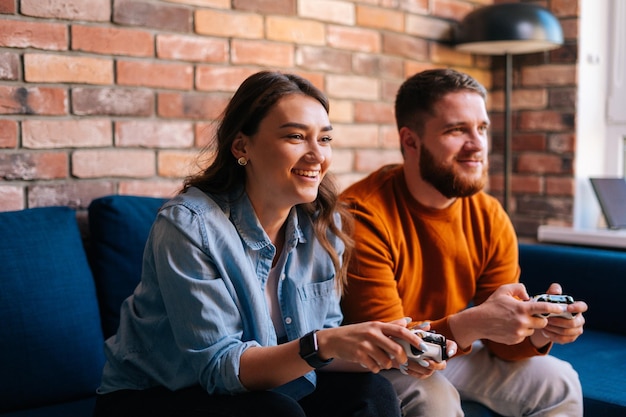 Felice coppia giovane allegra in possesso di controller e giocare ai videogiochi su console seduti insieme sul divano in un accogliente soggiorno. concetto di attività per il tempo libero degli amanti a casa.