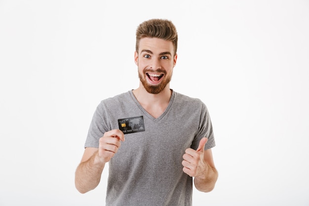 クレジットカードを持っている幸せな陽気な若い男は、親指を立てるジェスチャーをします。
