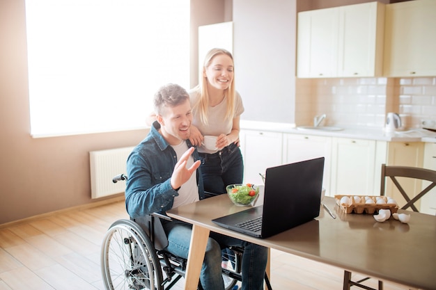 Happy cheerful young man sit at table and look on laptop. Guy with disability and inclusiveness. Young woman stand besides. Watching movie on lpatop.