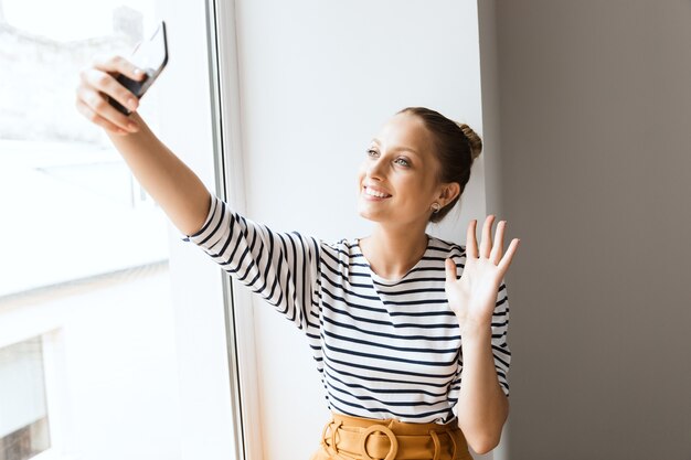 幸せな陽気な若いポジティブな女性は、携帯電話を振って話しているラップトップコンピューターを使用して屋内の窓の近くに座っています。