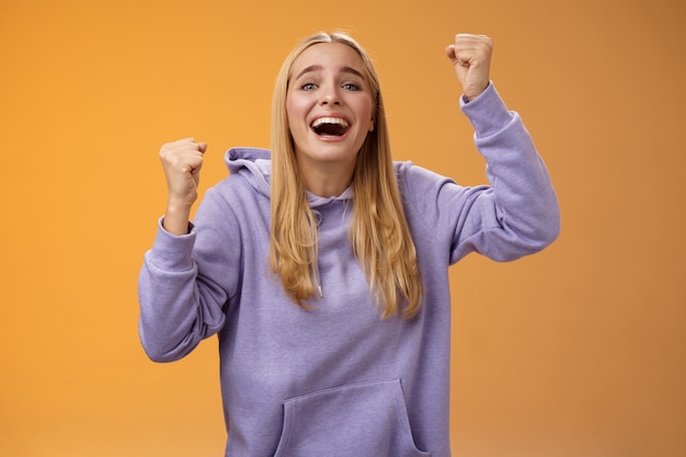 Photo happy cheerful young supportive woman cheering sister win smiling dedicated fun hope favorite team score goal raising clenched fists yelling happily celebrating triumphing victory, orange background.