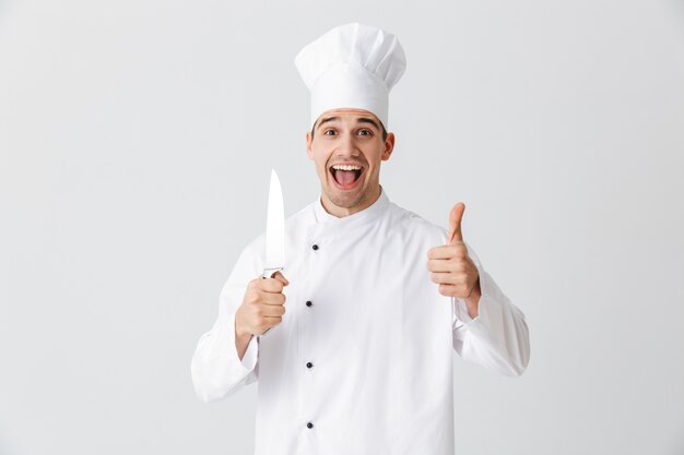 Happy chef cook wearing uniform holding a knife isolated over white wall