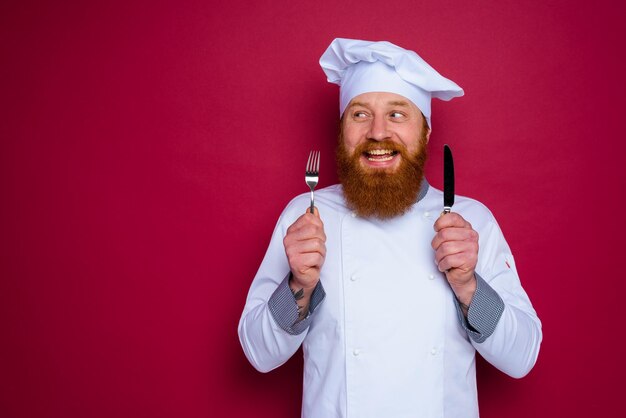 Happy chef with beard and red apron holds cutlery in hand