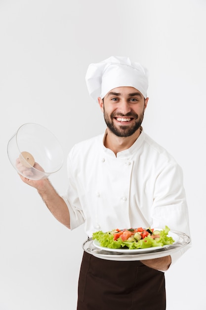 happy chief man in cook uniform smiling and holding plate with vegetable salad isolated over white wall