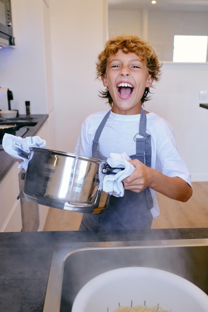 Happy child in apron with pot looking at camera against sink with pasta in colander at home