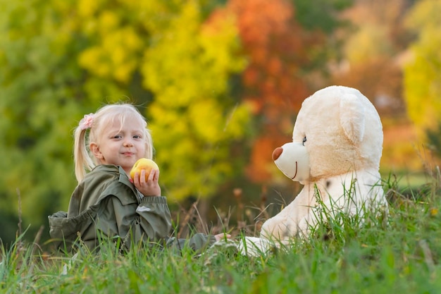 Bambino felice il giorno d'autunno in natura la bambina mangia la mela e gioca con il grande orsacchiotto seduto sull'erba