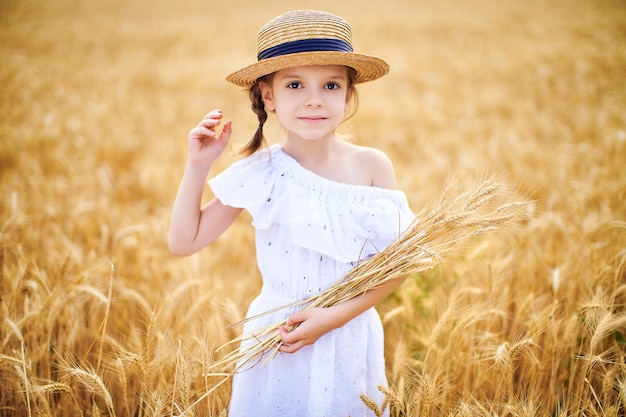 Happy child in autumn wheat field. Beautiful girl in white dress and straw hat have fun with playing, harvesting