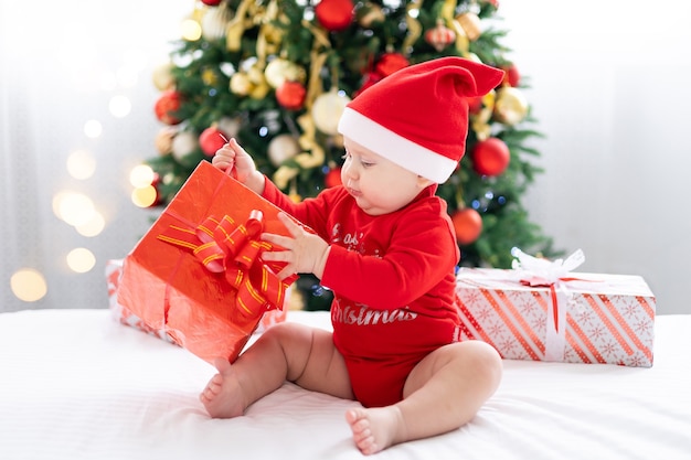 Happy child baby in red santa costume celebrating new year at home with christmas tree and gifts