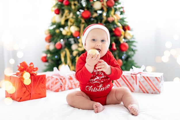 Happy child baby in red santa costume celebrating new year at home with christmas tree and gifts