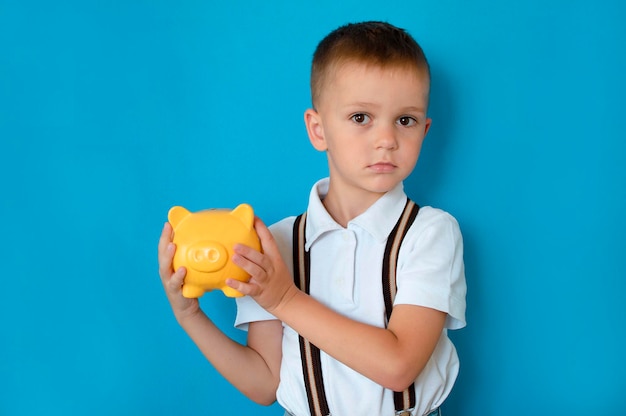 Happy child on blue background is saving money in piggy bank learning to save How to become rich Portrait of cute boy carrying piggy bank with his savings Concept of money insurance and people