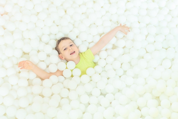 Happy child boy in the big dry pool with thousand of white balls Childhood concept Indoor leisure activity Having fun in playroom