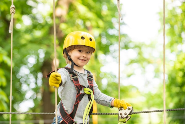 Счастливый ребенок мальчик звонит, взбираясь на высокое дерево и веревки. Портрет красивого ребенка в веревочном парке среди деревьев. Маленький мальчик наслаждается детскими годами.