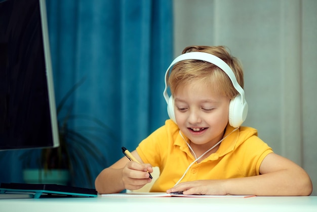 Счастливый ребенок мальчик в наушниках во время онлайн-обучения дома, делая домашнее задание