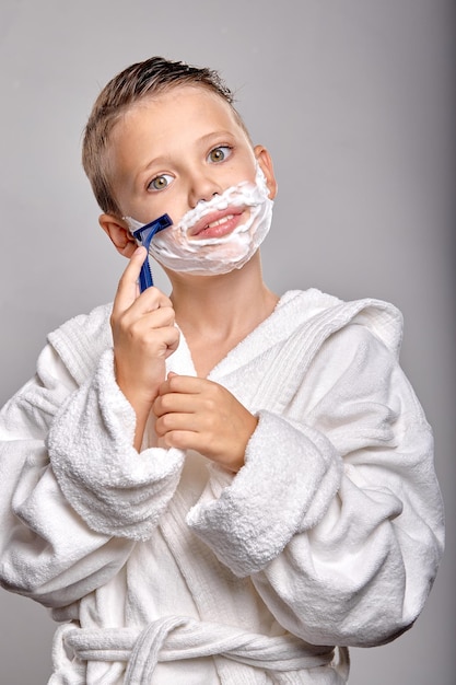 Счастливый мальчик-сын с приятной внешностью имеет пену для бритья на лице, держит бритву и собирается бриться, стоя перед зеркалом, изолированным на сером фоне. Молодой ребенок подражает отцу в халате