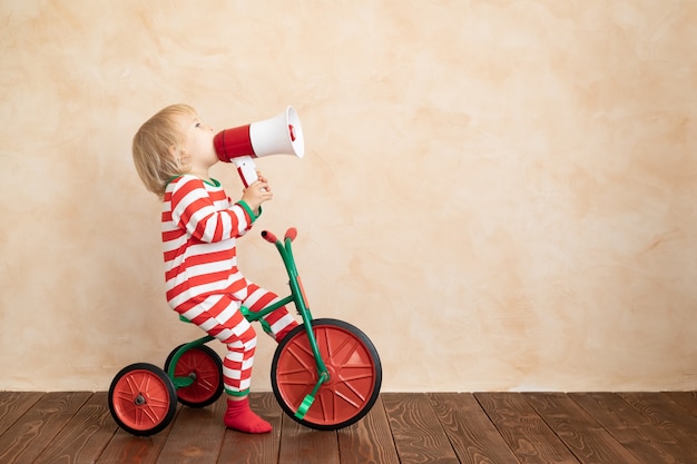 Happy child dressed Santa Claus costume playing at home. Funny kid driving toy car and speaking by megaphone. Christmas holiday concept