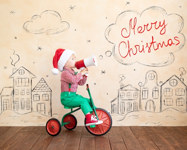 幸せな子供は家で遊んでいるサンタクロースの衣装を着ています。おもちゃの車を運転し、メガホンで話す面白い子供。クリスマス休暇のコンセプト