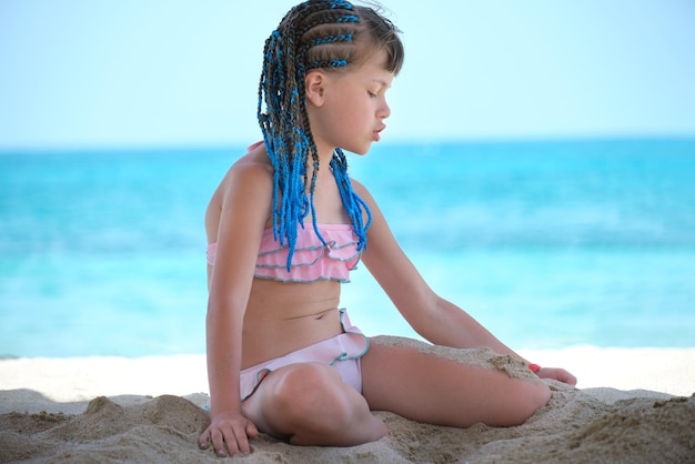 Happy child girl in bikini swimsuit playing with sand on seaside beach during summer tropical vacation