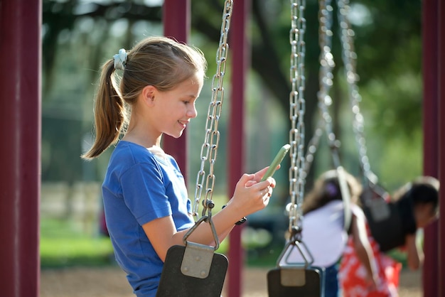 Bambina felice che naviga nel suo telefono cellulare seduto sull'altalena nel parco durante le vacanze estive