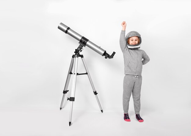 Happy child girl dressed in an astronaut costume standing beside the telescope.