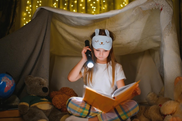 Photo happy child girl laughing and reading book in dark in a tent at home