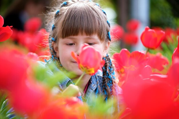 Happy child girl playing in summer garden enjoying sweet scent of red tulip flowers on sunny day