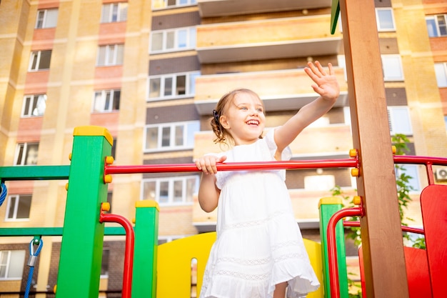 A happy child girl plays at the children's complex playground near the house in the summer in the yard and smiles