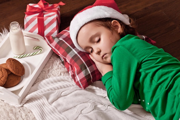 Happy child girl in Santa cap waiting Santa and sleep near pine tree on Christmas eve