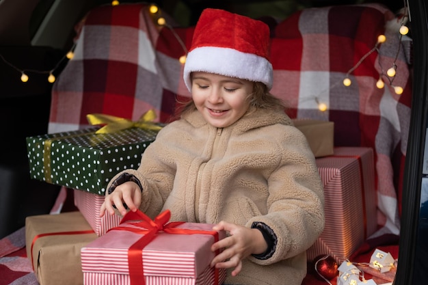 サンタの帽子をかぶった幸せな子供の女の子が車のトランクでクリスマス プレゼントを楽しむ