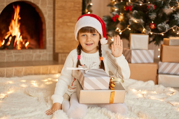 Happy child girl sitting near Christmas tree on Christmas Eve on floor and waving hand to camera