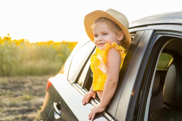 夕暮れ時の夏の自然の中で車の窓の外を見ている麦わら帽子の幸せな子の女の子