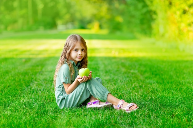 A happy child girl in the summer on the lawn with a green apple sits on the grass and smiles