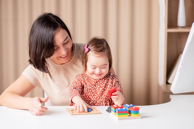 Bambina felice con sindrome di down con mamma con giocattoli educativi che il bambino al tavolo sta studiando