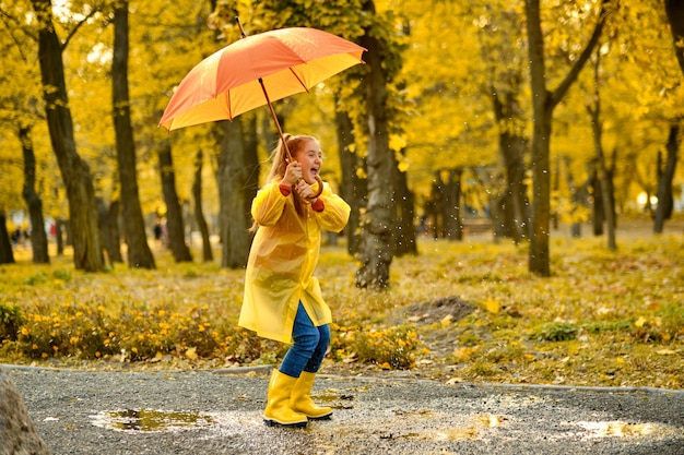 Bambina felice con un ombrello e stivali di gomma che saltano in una pozzanghera durante una passeggiata autunnale