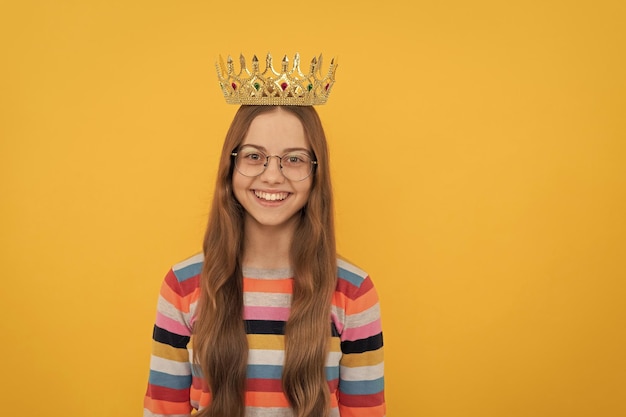 Happy child in glasses wear queen crown on yellow background victory