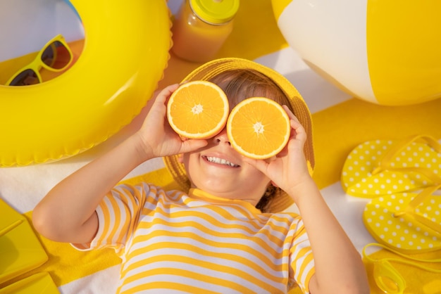 Happy child holding slices of orange fruit like sunglasses kid
wearing striped tshirt lying on beach towel healthy eating and
summer vacation concept