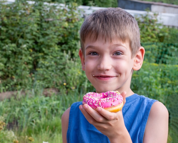 幸せな子供は公園でピンクのドーナツを食べています。不健康な食品の概念、甘い食べ物を間食。子供たちは甘い食べ物が大好きです。