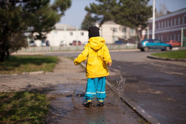 Bambino felice che salta in una pozzanghera in una tuta spray impermeabile in tutte le direzioni utili passeggiate per la città.