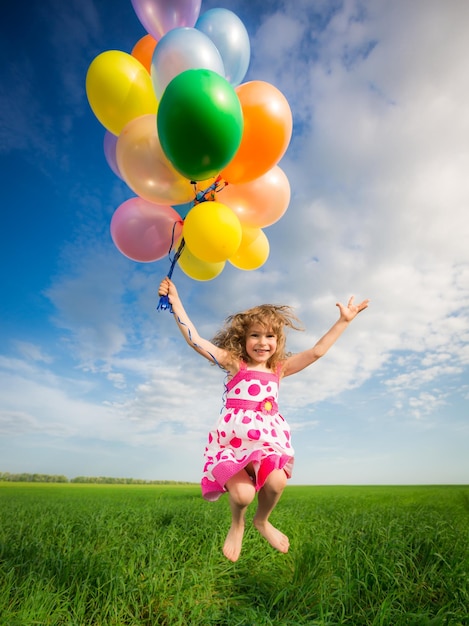 Happy child jumping with colorful toy balloons outdoors. Smiling kid having fun in green spring field against blue sky background. Freedom concept