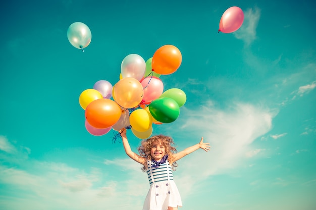 写真 カラフルなおもちゃの風船で屋外でジャンプする幸せな子。青い空を背景に緑の春のフィールドで楽しんで笑っている子供。自由の概念
