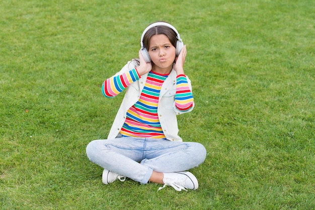 幸せな子供はヘッドフォンで音楽を聴きます。緑の芝生の上の小さな女の子。子供は公園の自然の中で歌を楽しみます。幸せな子供時代のコンセプト。現代生活でデジタル デバイスを使用します。女子高生は屋外でリラックスします。