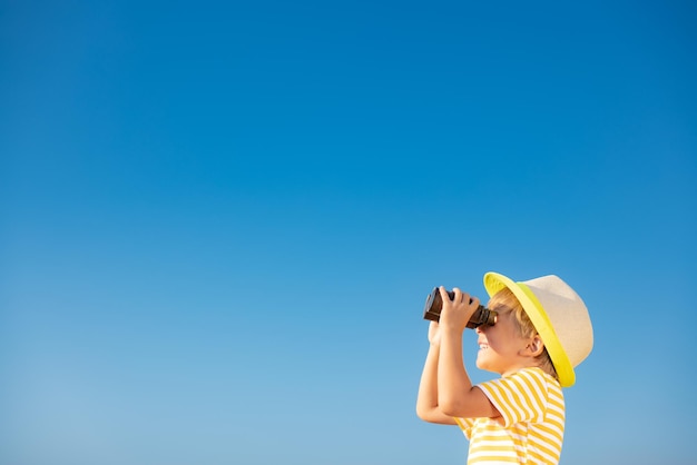 Happy child looking through binoculars against blue sky. Kid having fun outdoor in summer. Adventure and travel concept