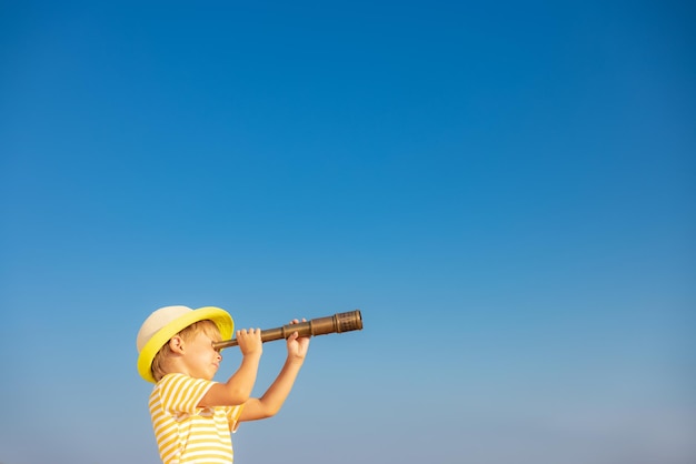 푸른 하늘을 배경으로 망원경을 통해 바라보는 행복한 아이 여름 모험과 여행 컨셉으로 야외에서 즐거운 시간을 보내는 아이