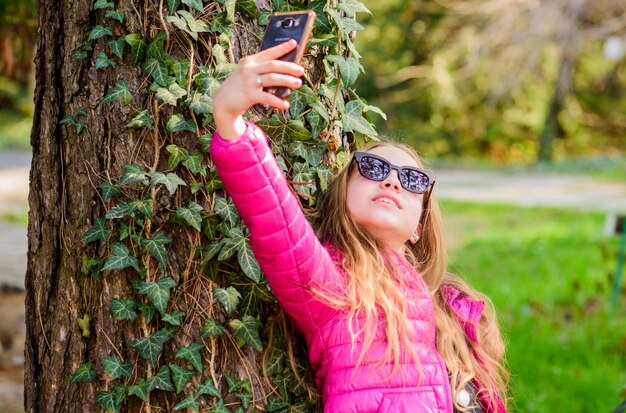 Happy child in park selfie time Natural beauty Childhood happiness summer nature little girl make selfie in park Spring holiday Green environment climbing plant on tree with ivy leaves