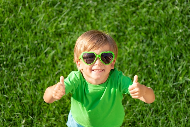 Happy child playing outdoor in spring park. Kid showing thumbs up on green grass. Earth day and ecology concept. Top view portrait of boy outside