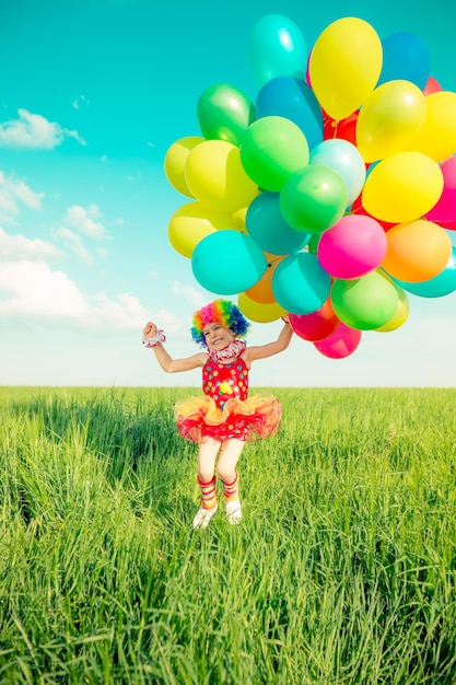 Happy child playing with colorful toy balloons outdoors. Smiling kid having fun in green spring field against blue sky background. Freedom concept