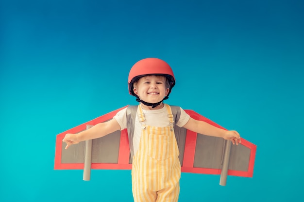 Happy child playing with toy paper wings against blue wall.