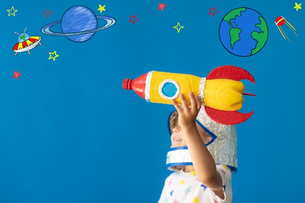 Happy child playing with toy rocket against blue background. Kid pretend to be astronaut. Imagination and children dream concept