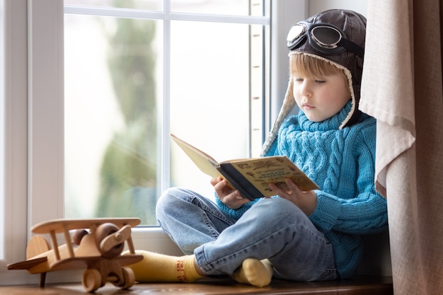 Happy child playing with vintage wooden airplane indoor. Kid reading book at home. Stay home and lockdown during coronavirus COVID-19 pandemic concept concept