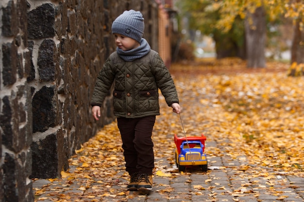 Happy child portrait with toy car on yellow autumn . Little smiling boy walking with big toy car in autumn city street and having fun